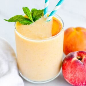 Homemade peach smoothie in a glass with straws and garnish next to fresh peaches.