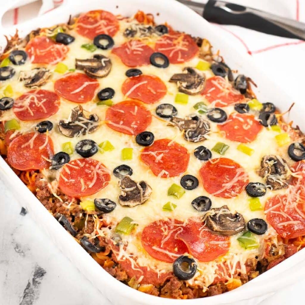 Baked pizza pasta in a white casserole dish topped with cheese, pepperonis, olives, and peppers.
