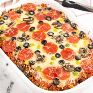 Baked pizza pasta in a white casserole dish topped with cheese, pepperonis, olives, and peppers.