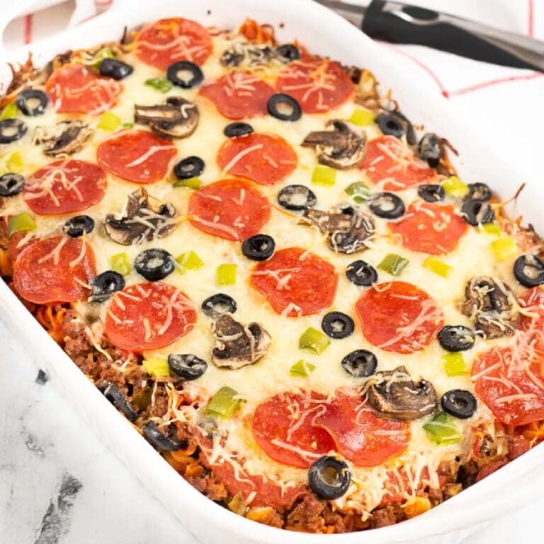 Baked pizza pasta in a white casserole dish topped with cheese, pepperonis, olives, and peppers.