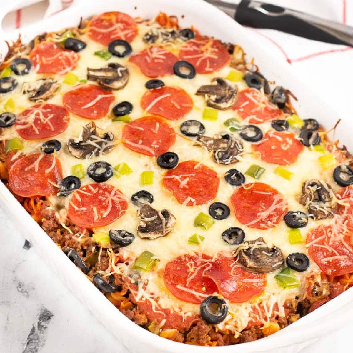 Baked pizza pasta in a white casserole dish topped with cheese, pepperonis, olives, and peppers.