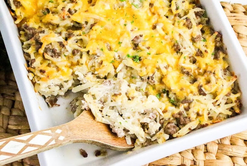 Scooping out a serving of hamburger and hash brown potato casserole from the dish with a wooden spoon.