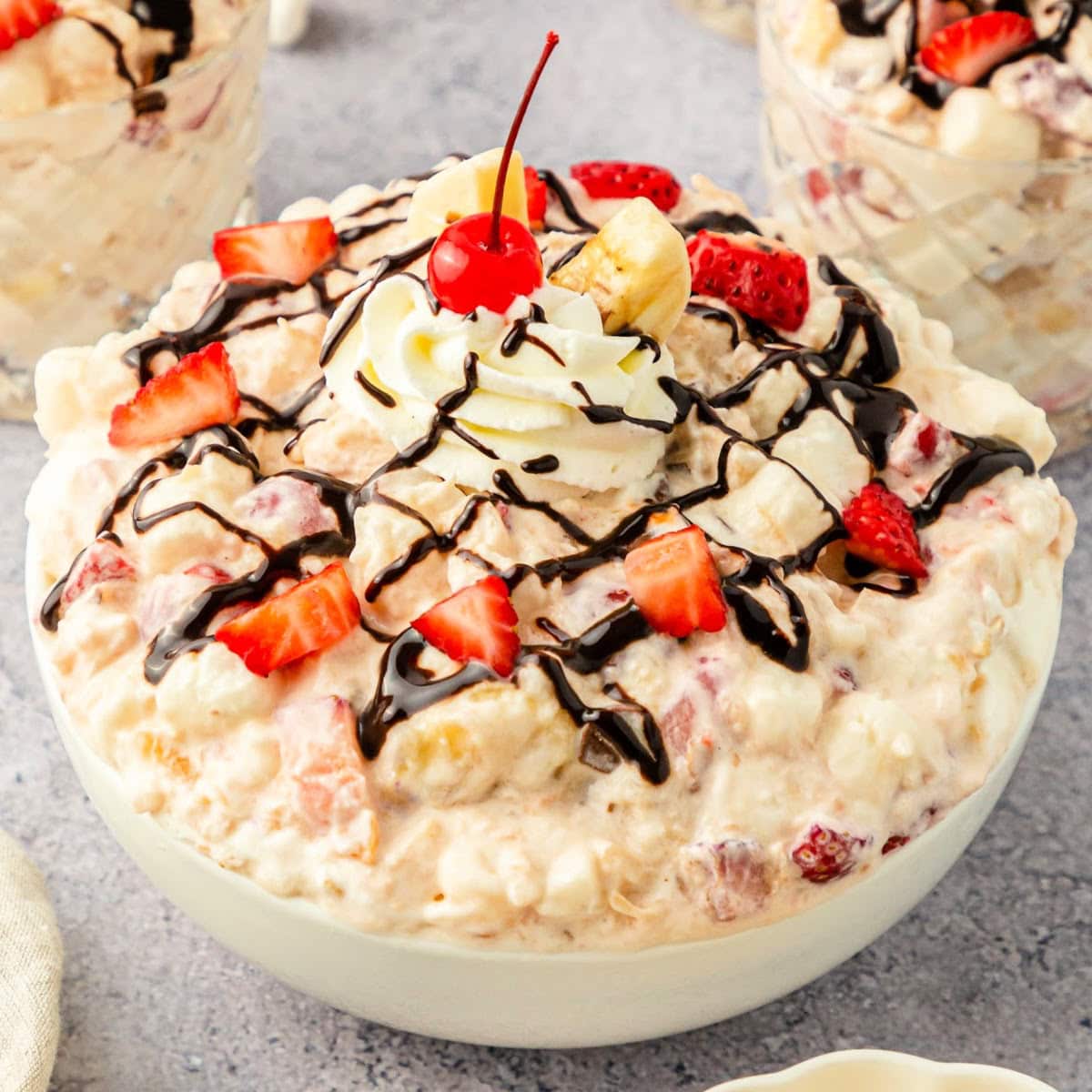 Banana split fluff salad topped with sliced strawberries, chocolate drizzle, whipped cream, and a cherrie.