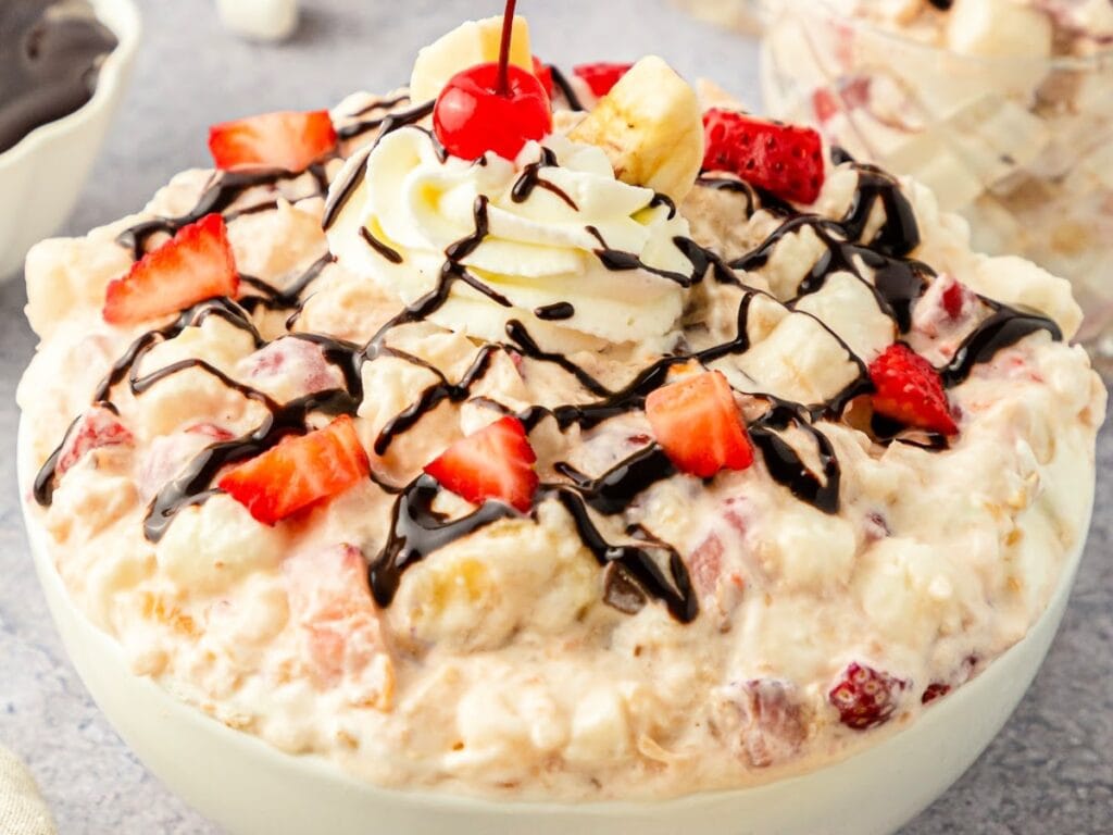 A large bowl of banana split salad topped with chocolate, strawberries, Cool Whip and a cherry.