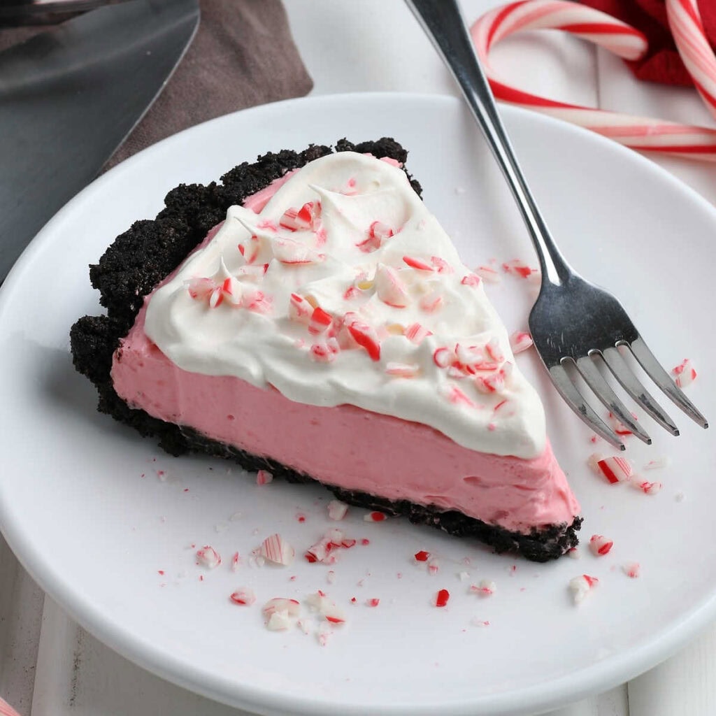 A slice of no bake peppermint pie garnished with whipped topping and crushed peppermints candies with a fork on a white plate.