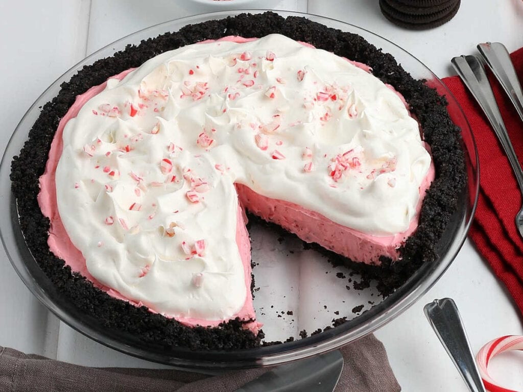 A no bake peppermint pie with a slice removed and garnished with whipped cream and crushed peppermint pieces.