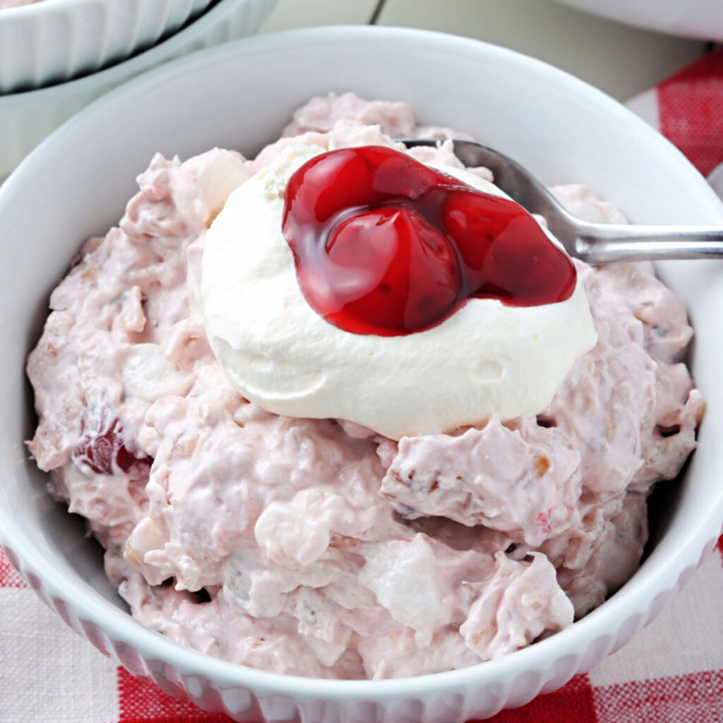Cherry fluff salad in a white bowl topped with a dollop of Cool Whip and a little cherry pie filling with a spoon.