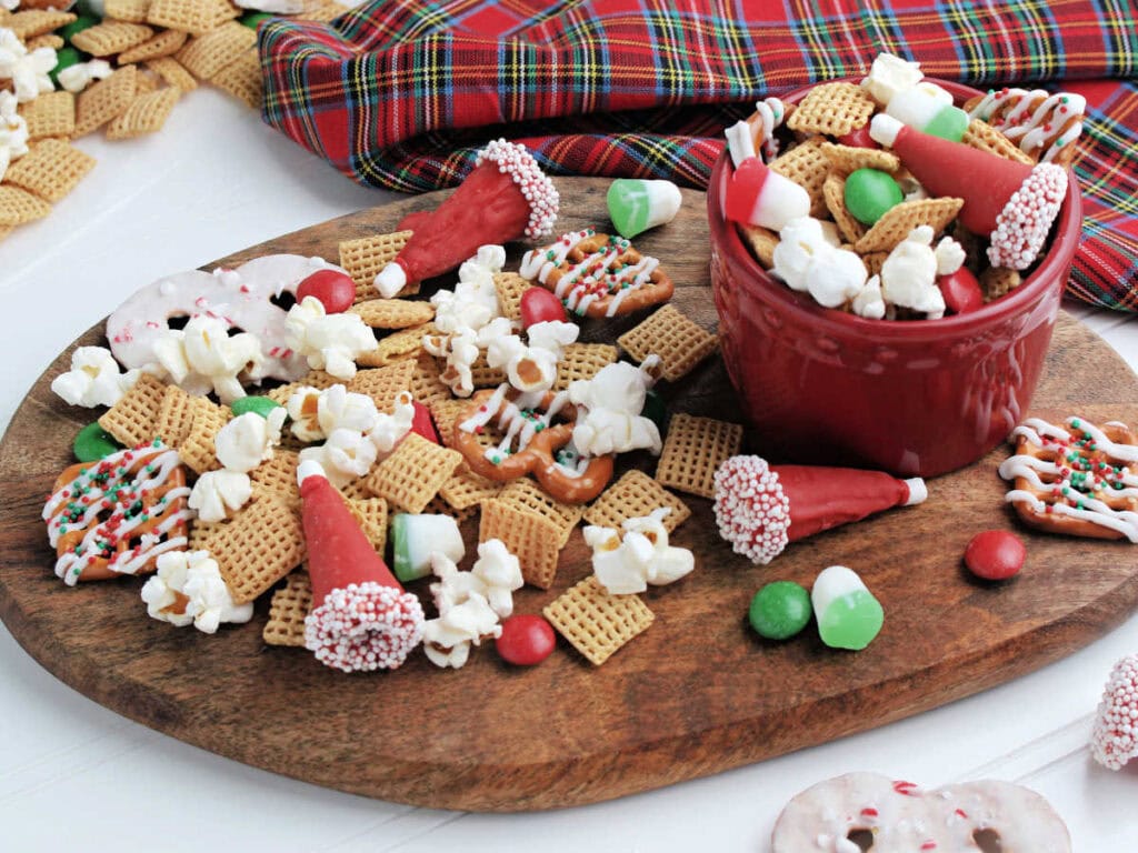 Holiday snack mix with pretzels in a red bowl on a wood cutting board.
