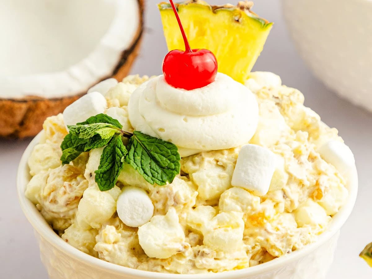Pineapple coconut fluff salad in a white bowl garnished with marshmallow, whipped topping, a cherry, and a pineapple wedge on top.