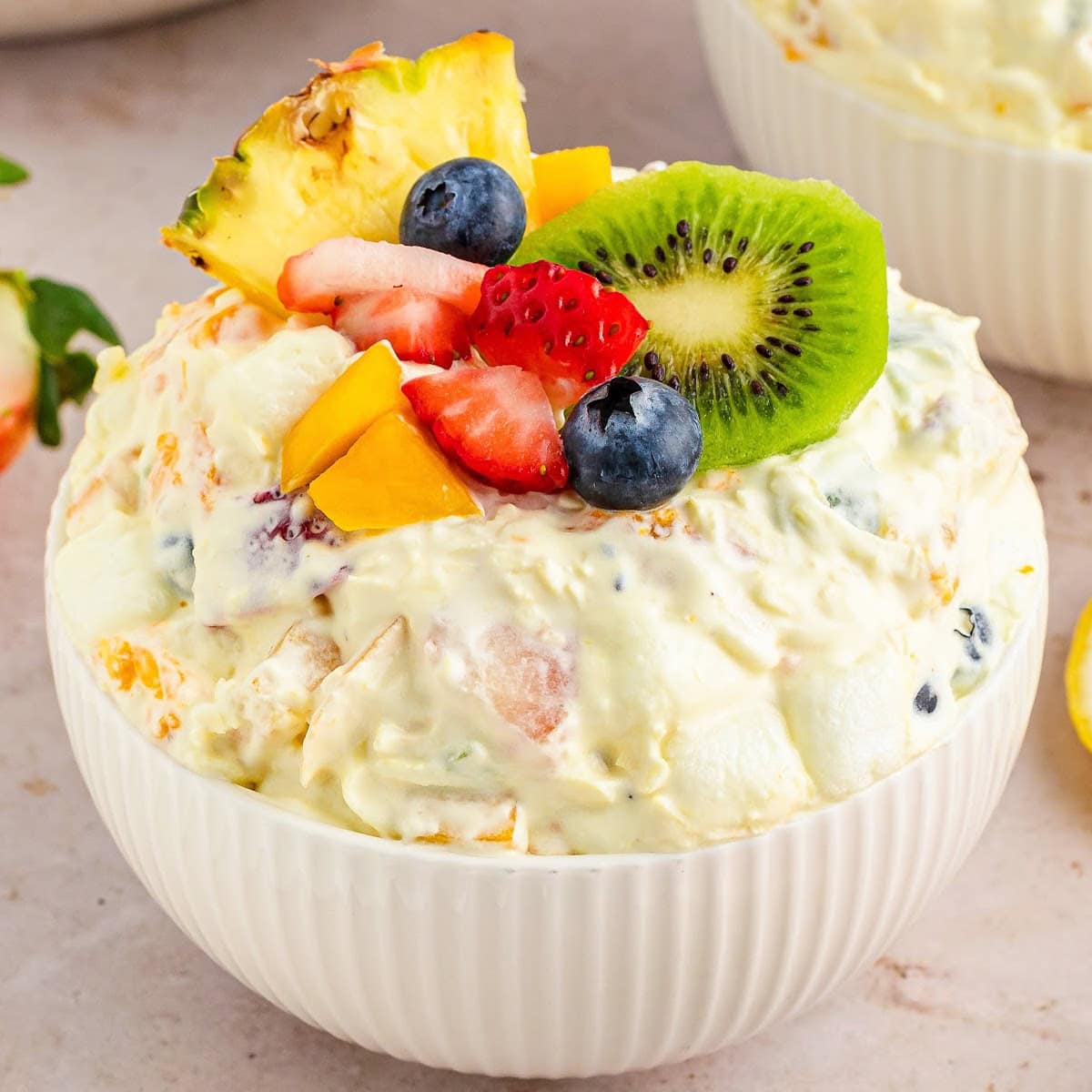 Tropical fruit fluff salad in a white bowl topped with kiwi, berries, and a pineapple wedge.