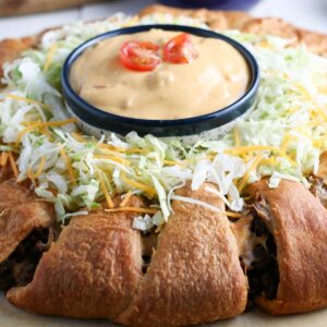 A taco crescent ring topped with lettuces and shredded cheese and a bowl of sauce in the middle.