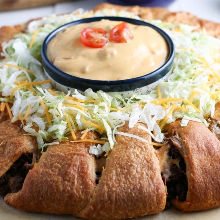 A taco crescent ring topped with lettuces and shredded cheese and a bowl of sauce in the middle.