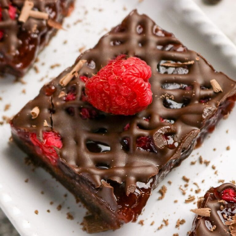 Chocolate raspberry bars on a white dish topped with a chocolate drizzle and fresh raspberry.