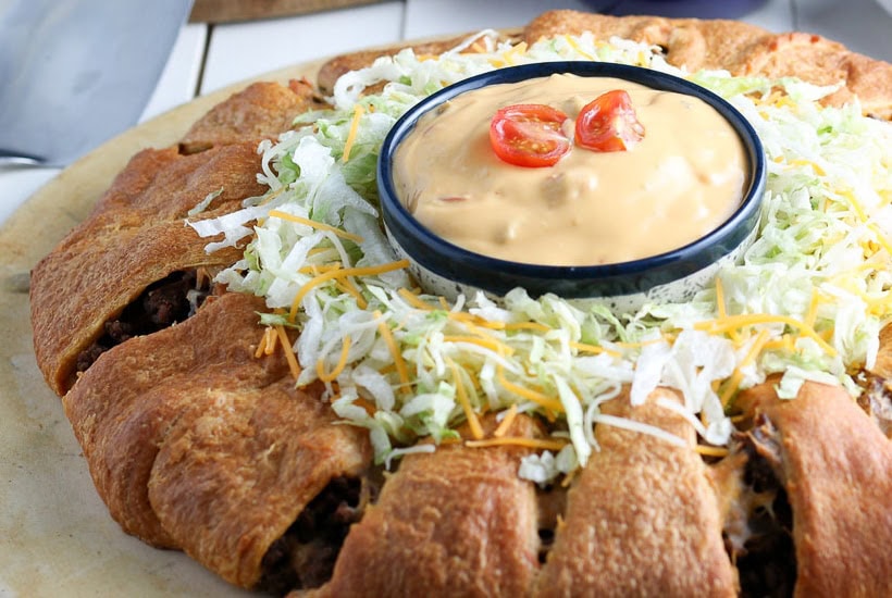 A crescent taco ring on a pizza stone garnished with lettuce and cheese on top with a ramekin of sauce in the middle.