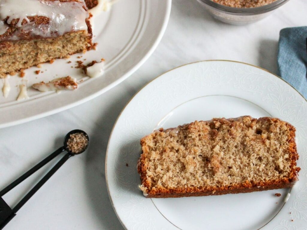 A slice of glazed cinnamon bread on a plate next to the loaf with icing on top.