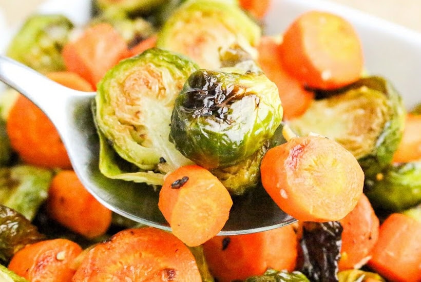 Scooping out baked brussels sprouts and carrots from a bowl using a spoon.
