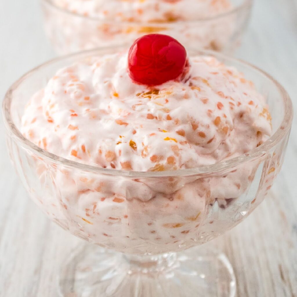 Acini de pepe fruit salad with a cherry on top in a glass bowl.