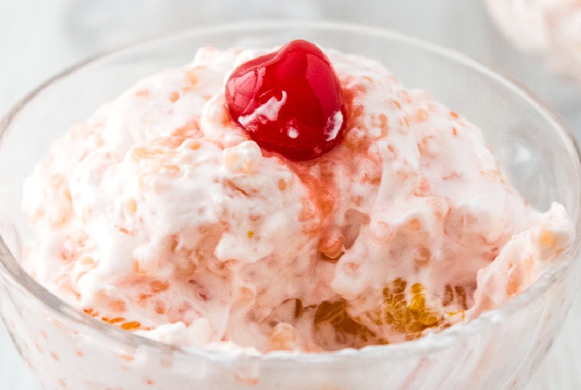 Acini de pepe fruit salad with pasta and Cool Whip garnished with a maraschino cherry in a glass serving bowl.