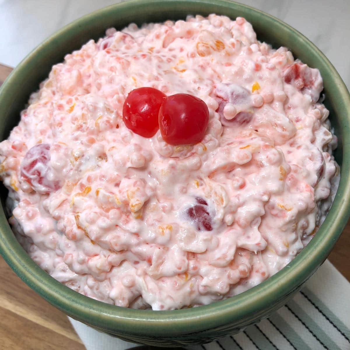 Green bowl filled with creamy acini de pepe fruit salad garnished with maraschino cherries.