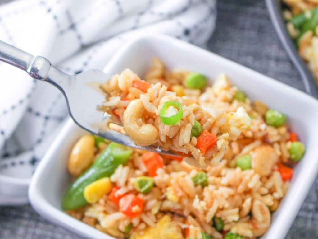 A forkful of fried rice with cashews rising over a bowl of rice.