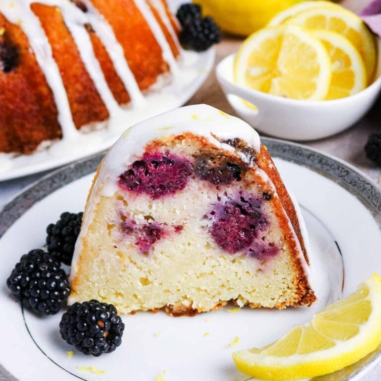 A slice of lemon blackberry bundt cake topped with lemon glaze on a white plate with a lemon slice and blackberries.