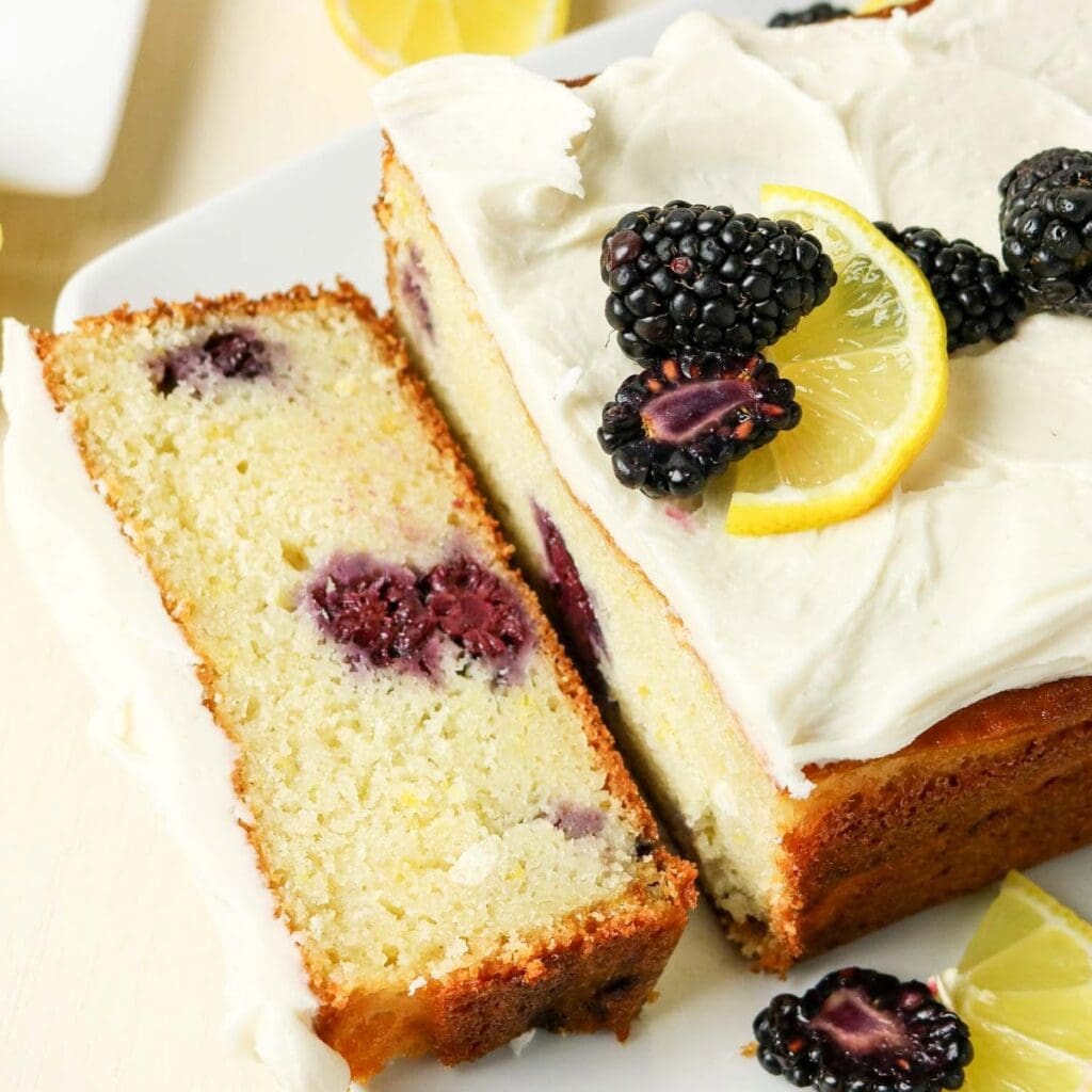 Frosted Lemon Blackberry Loaf with a slice cut off and garnished with lemon slices and berries on a cutting board.