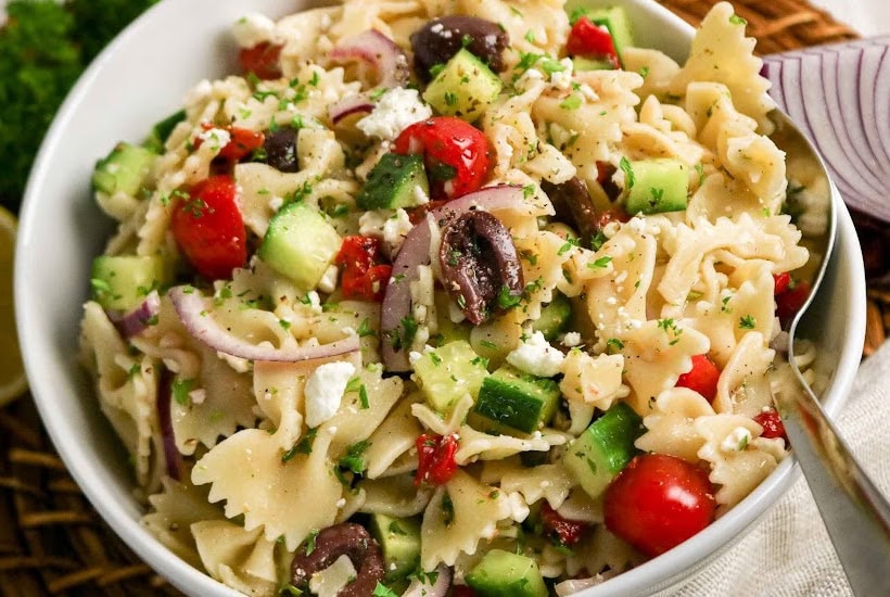 A white bowl full of pasta Mediterranean salad with a serving spoon.