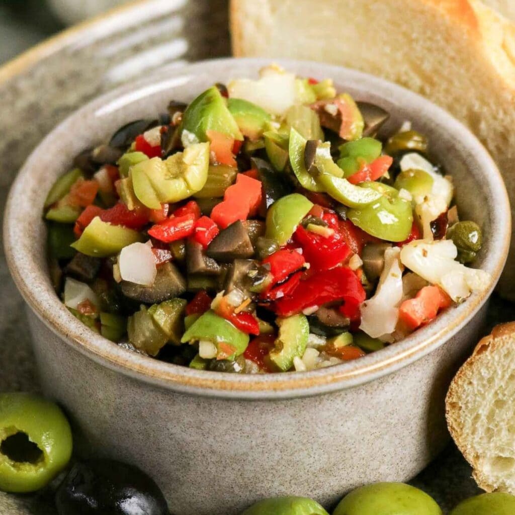 Small bowl filled with homemade Muffuletta Olive Salad with slices of bread and whole olives.