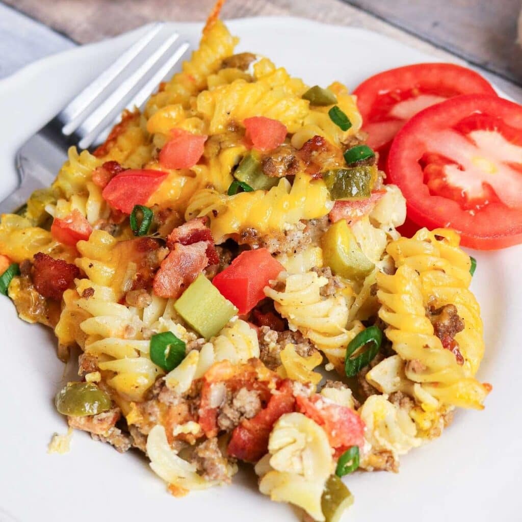 A serving of bacon cheeseburger casserole with tomato slices and a fork on a white plate.
