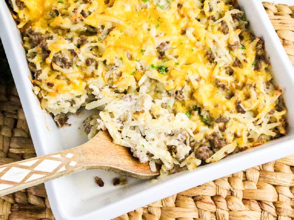 Scooping out a serving of hamburger hash brown casserole from the dish with a wooden spoon.