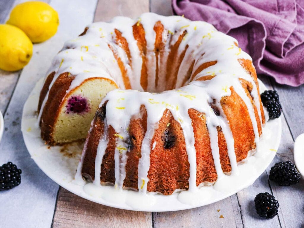 Lemon glazed blackberry bundt cake on a white serving platter surround by blackberries and lemons.