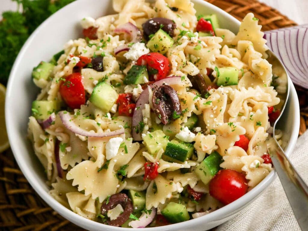A white bowl full of pasta Mediterranean salad with a serving spoon.