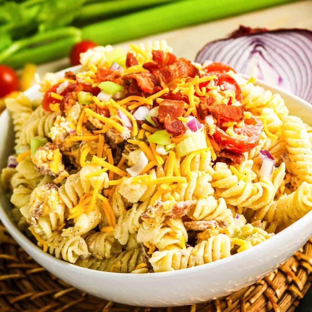 Closeup view of a round white round bowl filled with creamy pasta salad with bacon, cheese, red onion and tomatoes.