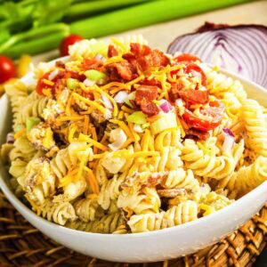 Closeup view of a round white round bowl filled with creamy pasta salad with bacon, cheese, red onion and tomatoes.