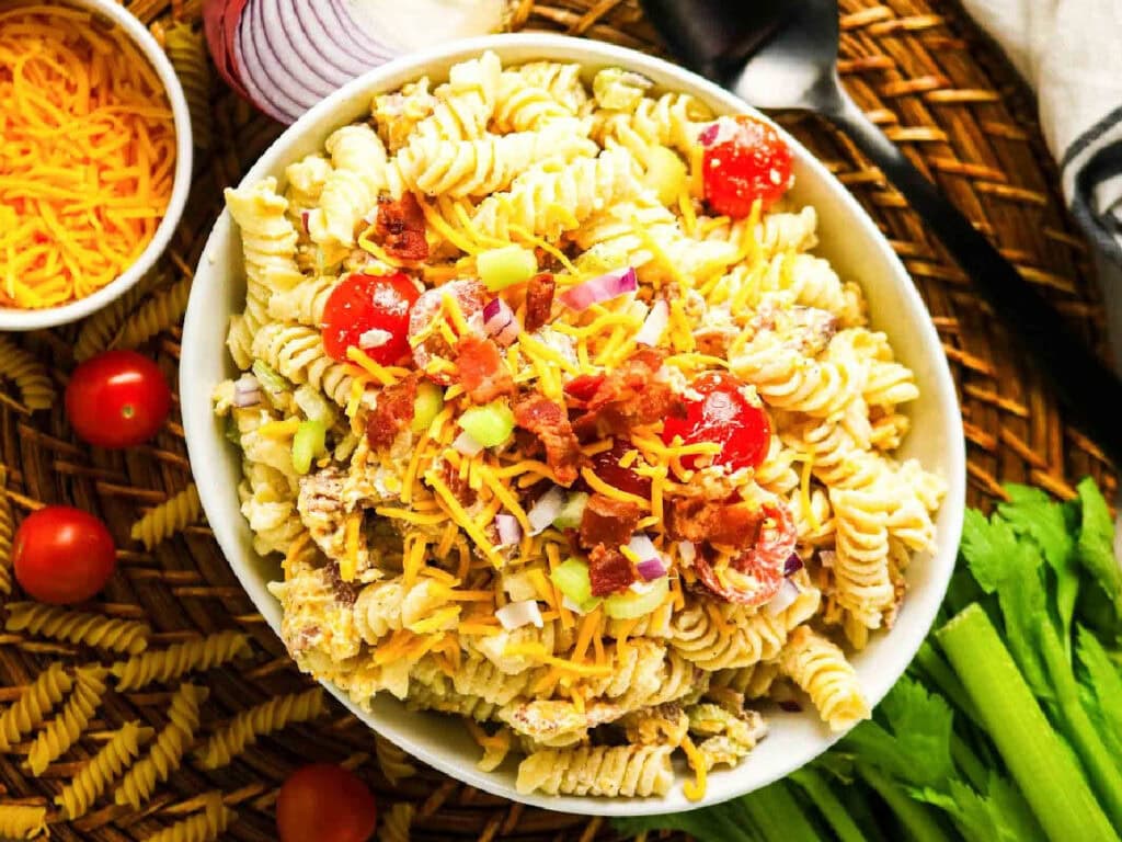 Creamy bacon pasta salad in a white serving bowl surrounded by miscellaneous ingredients.