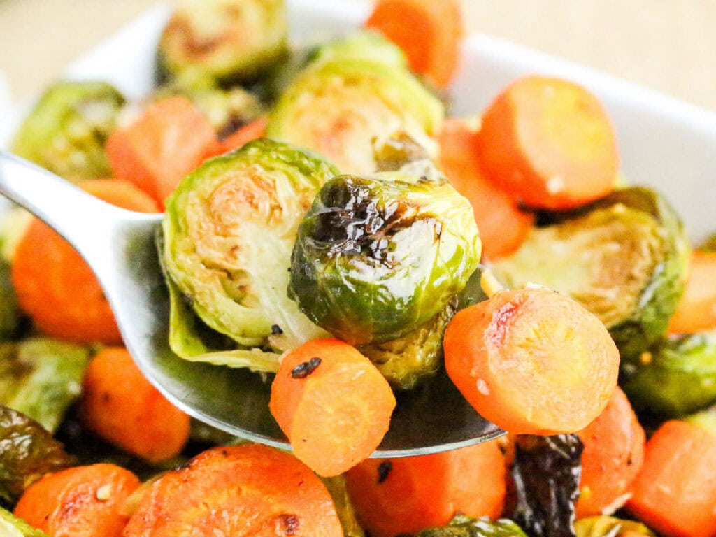 Scooping out baked brussels sprouts and carrots from a bowl using a spoon.