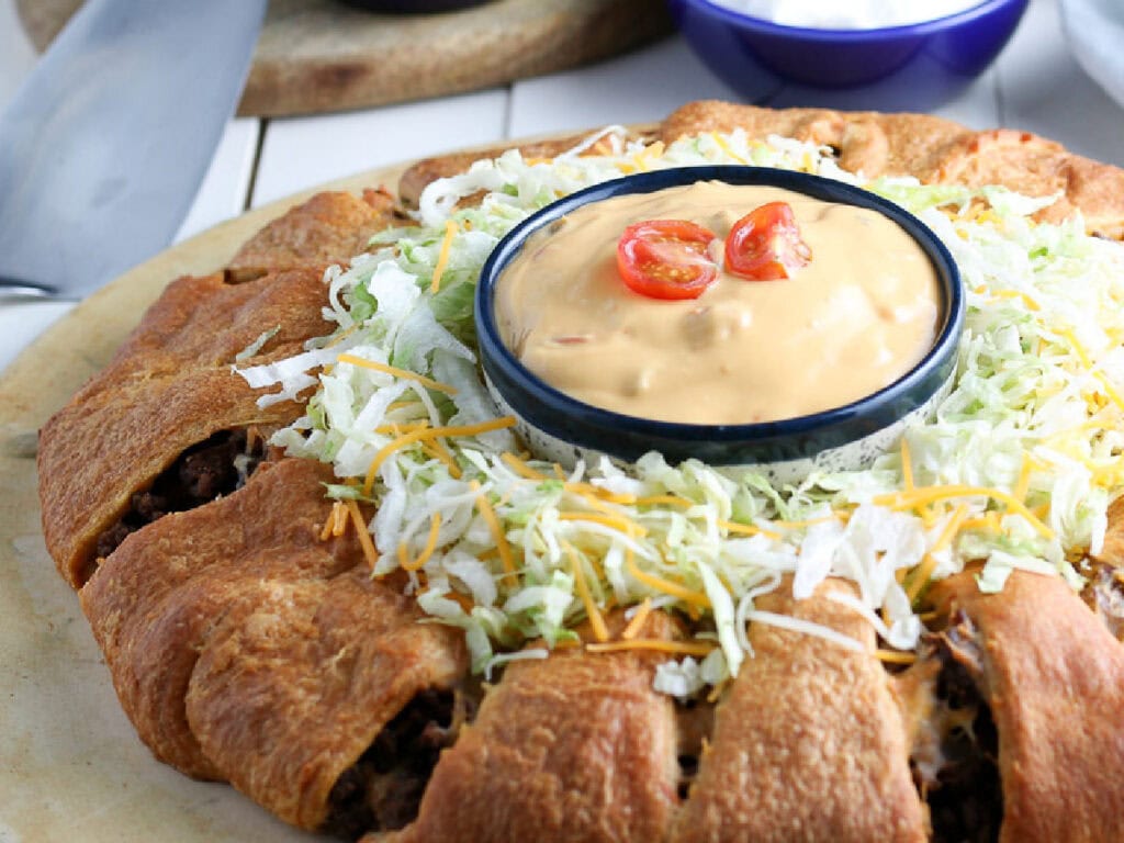 A crescent taco ring on a white plate with lettuce and cheese on top with a ramekin of sauce in the middle.