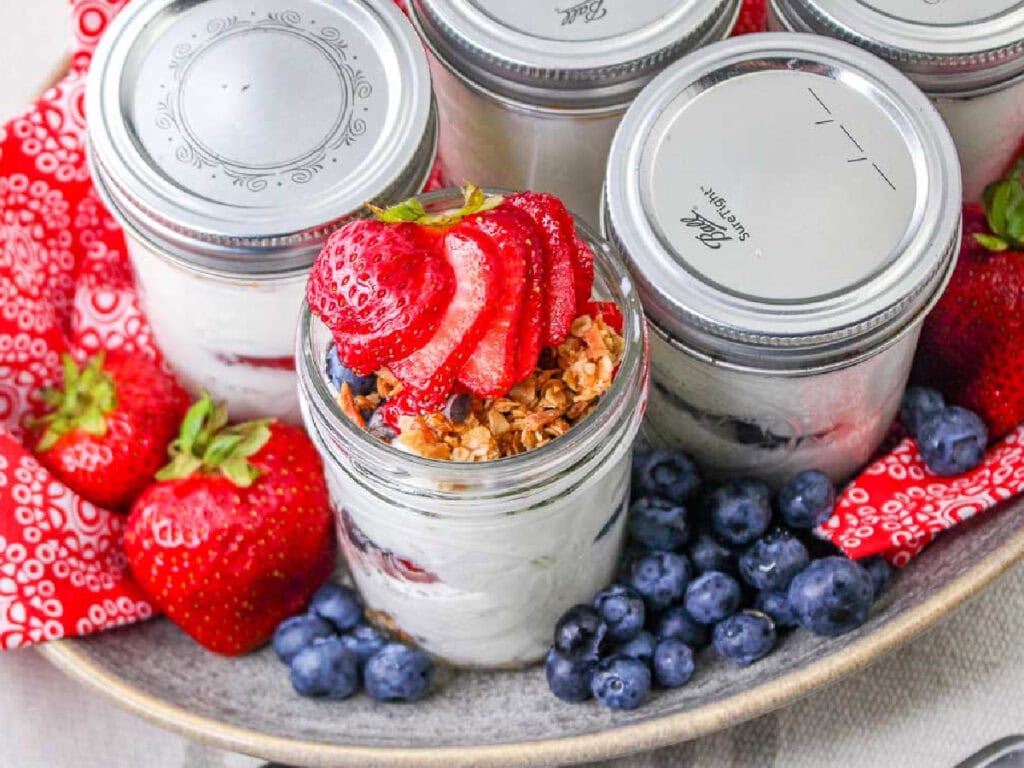 Greek yogurt parfaits topped with granola and strawberries in mason jars next to blueberries.